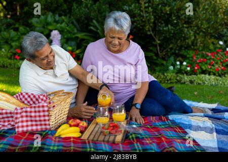 Homme biracial senior versant du jus dans le verre tout en appréciant pique-nique avec la femme contre les plantes dans le parc Banque D'Images