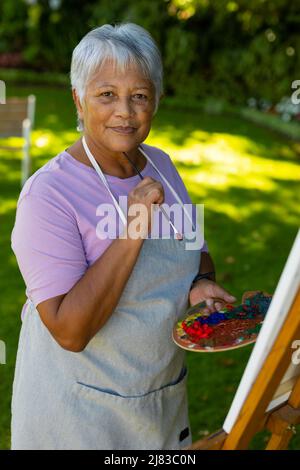 Portrait d'une femme biraciale âgée avec des cheveux courts portant un tablier tenant une brosse et une palette dans la cour Banque D'Images