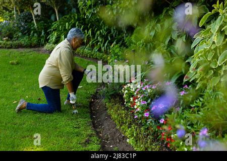 Vue latérale d'une femme biraciale senior avec jardinage de cheveux courts avec outil de fourche tout en s'agenouillant dans la cour Banque D'Images