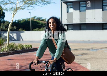 Portrait d'une jeune femme biraciale heureuse à vélo tout en se déplaçant en ville par beau temps