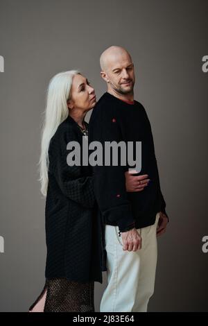 Vertical moyen long studio portrait de femme affectueuse mûre avec de longs cheveux gris qui embrasse l'homme de derrière, fond gris Banque D'Images