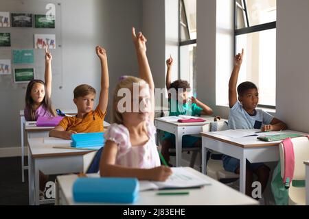 Les élèves de l'école primaire multiraciale se levant les mains tout en étant assis à leur bureau dans la salle de classe Banque D'Images