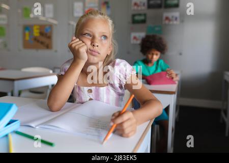 Écolière élémentaire de race blanche avec main sur le menton regardant loin tout en étudiant au bureau en classe Banque D'Images