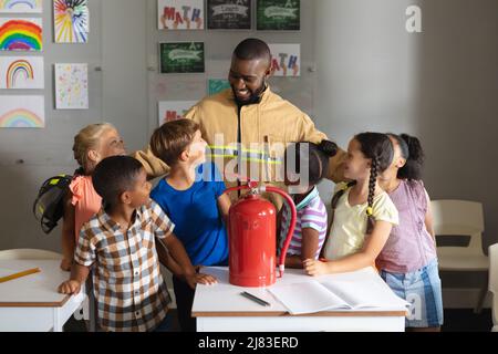 Étudiants élémentaires multiraciaux à la recherche d'un jeune homme afro-américain enseignant en uniforme de pompier Banque D'Images