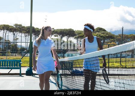 Souriant, de jeunes joueuses de tennis multiraciales parlent tout en marchant sur le court par beau temps Banque D'Images