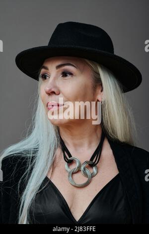 Portrait en gros plan vertical moyen d'une belle femme mûre avec de longs cheveux gris portant une tenue moderne de goth avec chapeau donnant sur l'extérieur Banque D'Images