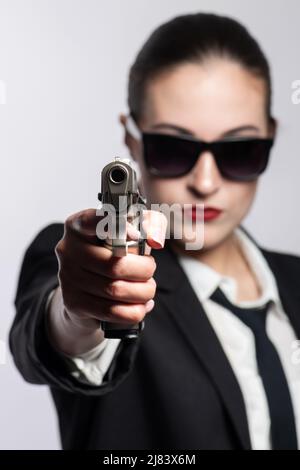 Belle et sérieuse femme avec des lunettes de soleil, costume noir classique, cravate et chemise blanche pointant pistolet à l'appareil photo. Modèle avec rouge à lèvres. Pistolet b Banque D'Images