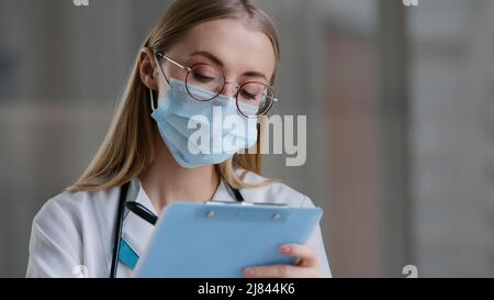 Portrait femme caucasienne médecin infirmière dans le masque de protection médicale écriture de l'information dans le journal d'enregistrement médical femme dans lunettes thérapeute Banque D'Images