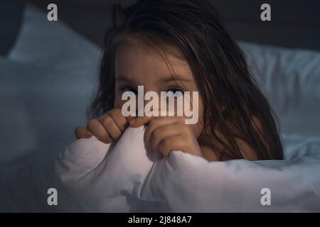 Une petite fille mignonne se couche dans le lit, effrayée et couvre son visage avec une couverture. Enfants. Photo de haute qualité Banque D'Images