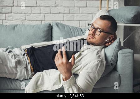 Homme caucasien couché sur le canapé à la maison et écoutant de la musique au téléphone. Communication Internet, technologie de connexion sans fil. Mobile Banque D'Images