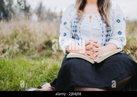 Femme méconnaissable assise à l'extérieur sur l'herbe priant avec ses mains sur une Bible ouverte. Banque D'Images