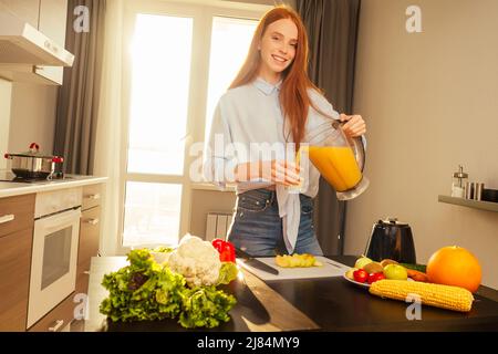 Belle femme au gingembre aux cheveux rouges qui fait des smoothies de fruits avec un mélangeur, préparant la boisson avec des bananes, de la pomme et de l'orange à la maison dans la cuisine Banque D'Images