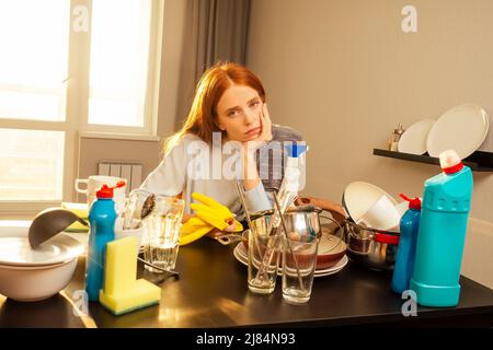 malheureux gingembre rougeâtre belle femme portant le gant jaune et la pile de lavage montagnes de plats sales: tasses, poêle, casserole dans la cuisine Banque D'Images