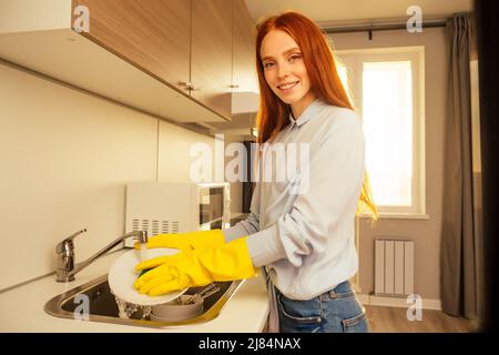 malheureux gingembre rougeâtre belle femme portant le gant jaune et la pile de lavage montagnes de plats sales: tasses, poêle, casserole dans la cuisine Banque D'Images