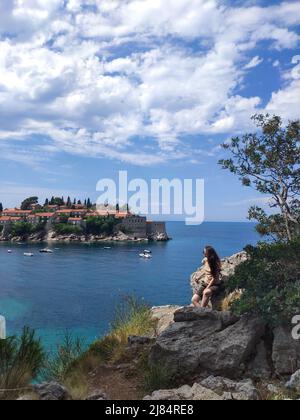 Une jeune femme est assise sur le rocher et regarde une belle vue sur l'île de Sveti Stefan au Monténégro pendant la journée Banque D'Images