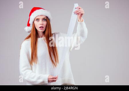 femme au gingembre aux cheveux rouges et surprise portant un chapeau de père noël rouge et un pull tenant une longue liste de reçus de shopping en studio blanc Banque D'Images