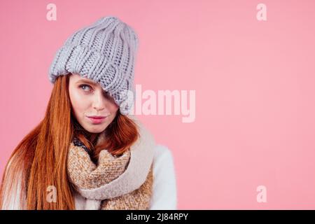 timide redhead ginger femme mettant un chapeau tricoté sur sa tête, cachant l'oeil et la bouche ouverte dans le fond rose studio Banque D'Images