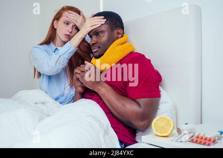 homme afro-américain en écharpe tricotée et femme au gingembre aux cheveux rouges en pyjama allongé dans le lit jour de congé du travail parce qu'ils se sentent de mauvaise humeur Banque D'Images