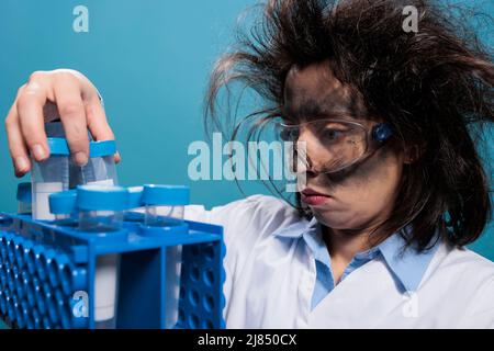 Un scientifique fou avec un visage sale et des cheveux désordonnés analysant des flacons de test en plastique après une explosion chimique de laboratoire. Chimiste fou ayant un rack avec des tubes à essai tout en essayant d'identifier le bon composé. Banque D'Images