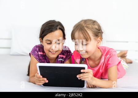 Deux petites filles mignonnes utilisent une tablette numérique et sourient lorsqu'elles sont allongées sur le lit dans la chambre des enfants Banque D'Images