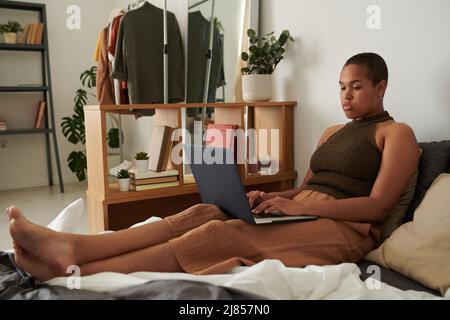 Jeune femme noire sérieuse assise sur le lit et dactylographiant sur un ordinateur portable tout en travaillant sur le projet marketing à distance pendant la quarantaine Banque D'Images