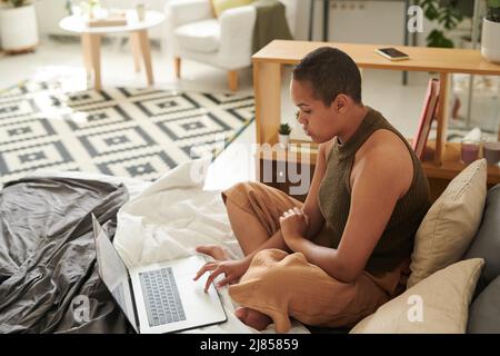 Jeune femme noire concentrée avec buzz coupé assis sur le lit et utilisant le pavé tactile d'ordinateur portable dans le studio plat Banque D'Images