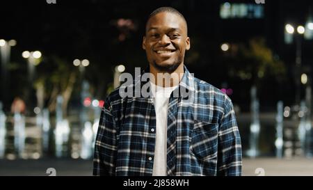 Portrait afro-américain 20s millénaire satisfait homme beau gars touriste se tient dans la ville en arrière-plan de soirée regardant l'appareil-photo souriant Toothy Banque D'Images