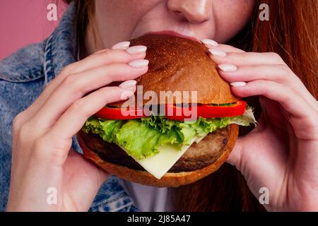 photo rognée d'une femme rouge au gingembre et aux cheveux rouges mangeant un gros cheeseburger avec du fromage, des côtelettes de bœuf, de la laitue tomate et des doigts de lécher de près Banque D'Images