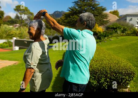 Joyeux mari aîné biracial dansant avec une femme senior sur une colline herbeuse dans le parc pendant la journée ensoleillée Banque D'Images