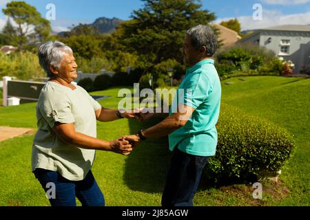 Vue latérale d'un heureux couple biracial senior se tenant les mains et dansant dans le parc pendant la journée ensoleillée Banque D'Images