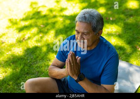 Vue en grand angle de l'homme aîné biracial avec les yeux fermés méditant tout en étant assis sur le tapis au-dessus de l'herbe Banque D'Images