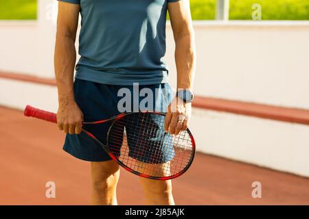 Section médiane de l'homme aîné biracial portant des vêtements de sport tenant une raquette pendant qu'il se tient debout sur le terrain Banque D'Images