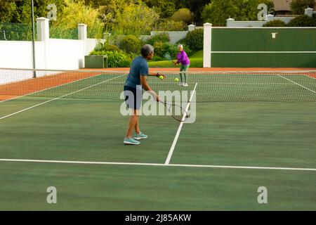 Pleine longueur de biracial homme senior servant le ballon à la femme senior tout en jouant au tennis dans le court Banque D'Images