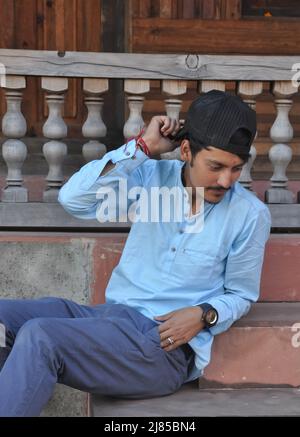 Vue latérale d'un jeune homme du nord indien qui regarde bien en touchant sa casquette en regardant vers le bas tout en étant assis sur les escaliers du temple Banque D'Images