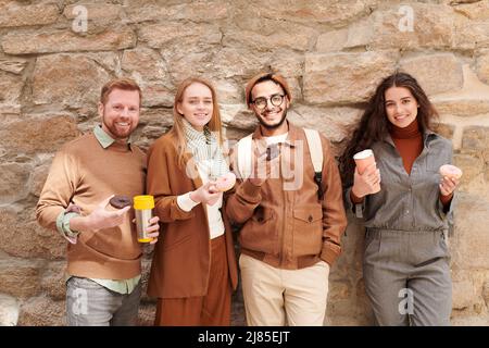 Portrait de jeunes amis multiethniques à la mode positifs voyageant ensemble debout avec des tasses de café et des beignets contre le vieux mur Banque D'Images