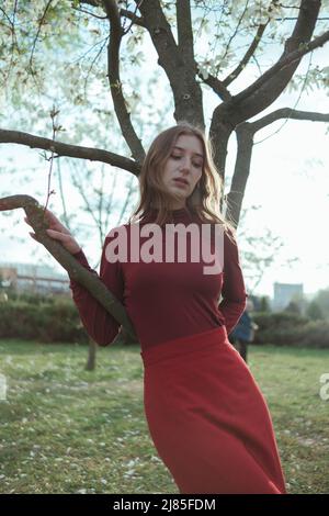 Une blonde en rouge s'appuie sur un arbre dans le parc. Le concept d'unité avec la nature Banque D'Images