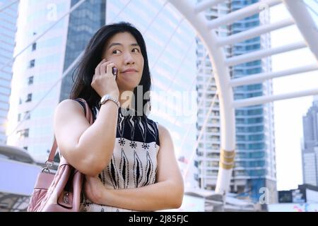 Une jeune femme d'affaires asiatique attirante en robe blanche fait un visage tout en parlant au téléphone, se sentant douteuse et sceptique. Banque D'Images