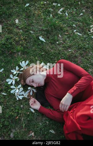 une blonde en rouge se trouve au printemps sur l'herbe entourée de fleurs magnolias Banque D'Images