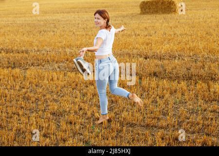 une fille pieds nus avec des baskets à la main marchant dans le champ agricole avec des botte de foin et des balles après la récolte. Banque D'Images