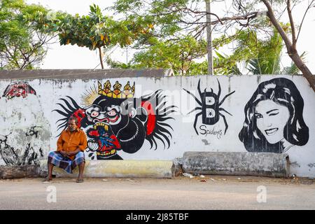 Vieil homme assis devant un mur couvert de graffiti à Tangassery, Thangassery, Kerala, Inde. Banque D'Images