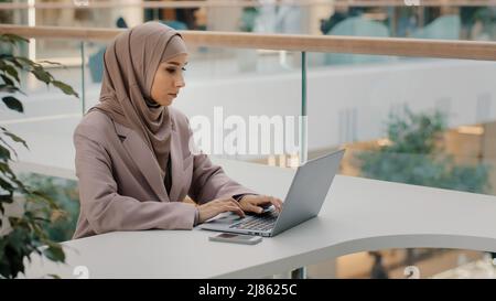 Concentrée sérieuse arabe femme d'affaires professionnelle entrepreneur assis au bureau dactylographie regardant l'écran d'ordinateur portable étudiant cours en ligne musulmane fille Banque D'Images