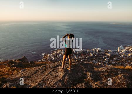 Femme en vêtements de sport debout sur la falaise et regardant la vue au coucher du soleil. Femme de randonnée se détendant sur le sommet de la montagne en regardant l'océan. Banque D'Images