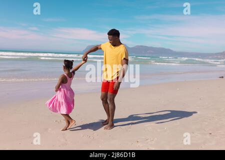 Joyeux jeune africain américain dansant avec sa fille à la plage contre la mer et le ciel le jour ensoleillé Banque D'Images