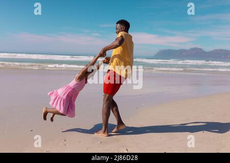 Jeune Afro-américain tenant les mains de sa fille et la faisant tourner à la plage contre le ciel bleu Banque D'Images