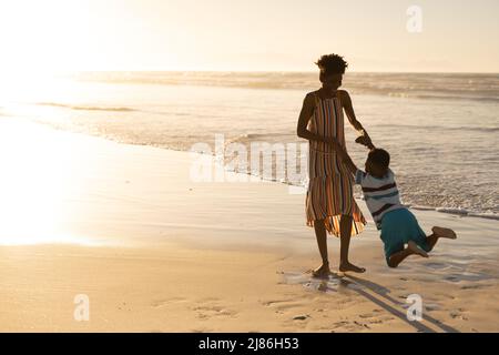 Joueuse jeune femme afro-américaine tenant les mains de son fils et lui faisant tourner à la plage contre le ciel Banque D'Images