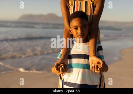 Portrait d'un mignon garçon afro-américain tenant les mains de la jeune mère tout en se tenant contre la mer Banque D'Images