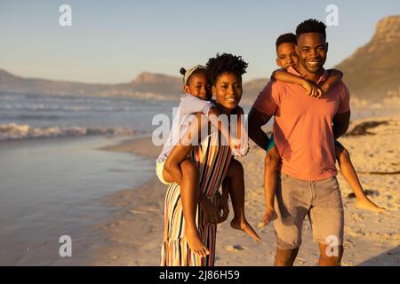 Portrait de jeunes parents afro-américains heureux, coggysoutenant leur fils et leur fille à la plage contre le ciel Banque D'Images