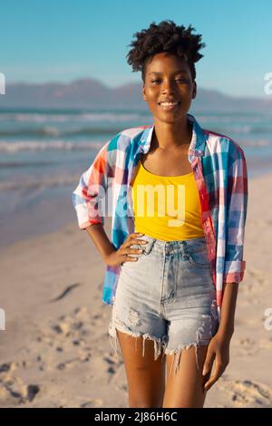 Portrait d'une jeune femme afro-américaine souriante aux cheveux courts debout à la plage contre le ciel bleu Banque D'Images