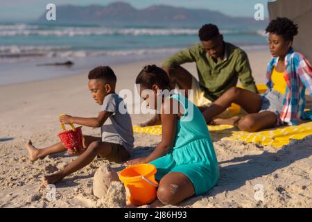 Jeunes parents afro-américains assis avec leur fils et leur fille jouant avec des seaux et du sable à la plage Banque D'Images