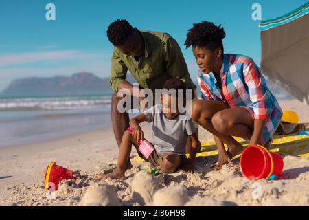 Jeunes parents afro-américains regardant son fils jouer avec du sable à la plage contre le ciel par beau temps Banque D'Images